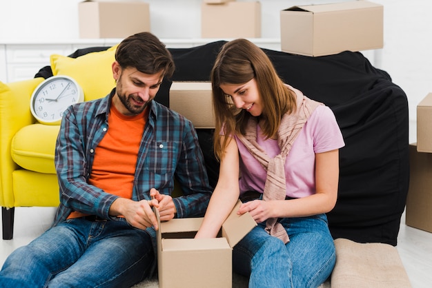 Pareja joven sonriente que se sienta cerca del sofá que desempaqueta la caja en nueva casa