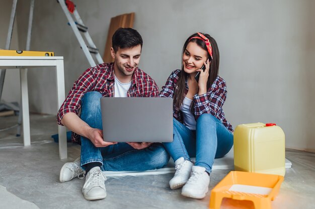 Pareja joven sonriente renovando y remodelando su nuevo apartamento, el hombre está pintando las paredes con un rodillo y la mujer se está conectando con una computadora portátil