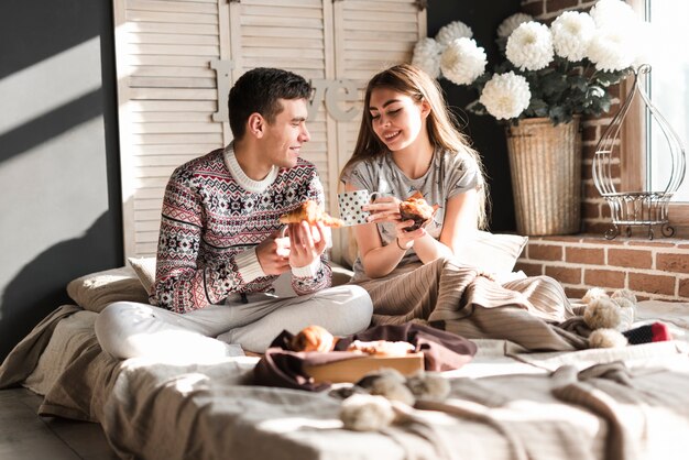 Pareja joven sonriente sentada en la cama con croissant y cupcake en la mano