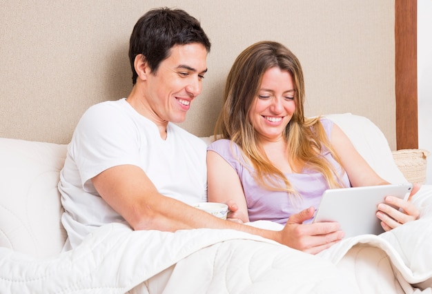 Pareja joven sonriente sentada en la cama mirando tableta digital