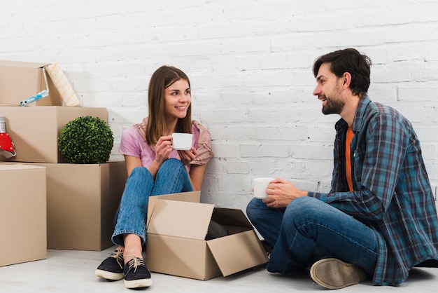 Pareja joven sonriente sentada en el piso con cajas de cartón en movimiento tomando el café