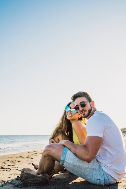 Foto gratuita pareja joven sonriente sentada por la playa