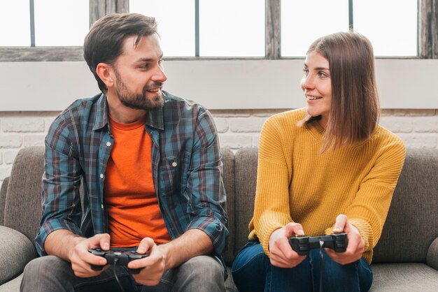 Pareja joven sonriente sentada en el sofá mirando el uno al otro mientras juega videojuegos