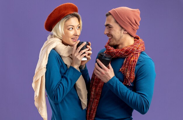 Pareja joven sonriente con sombrero con bufanda en el día de San Valentín mirando el uno al otro sosteniendo una taza de café aislado sobre fondo azul.