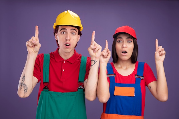 Pareja joven sorprendida en uniforme de trabajador de la construcción chico con casco de seguridad chica con gorra mirando a cámara apuntando hacia arriba aislado en la pared púrpura