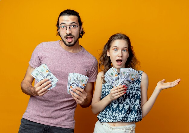 Pareja joven sorprendido mostrando dinero en efectivo de pie sobre la pared naranja