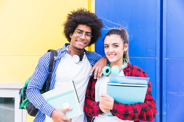 Pareja joven sosteniendo libros en la mano de pie contra la pared azul y amarilla