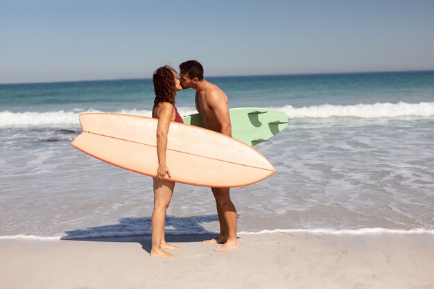 Pareja joven con tabla de surf besándose en la playa bajo el sol