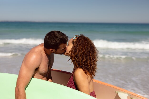 Pareja joven con tabla de surf besándose en la playa bajo el sol