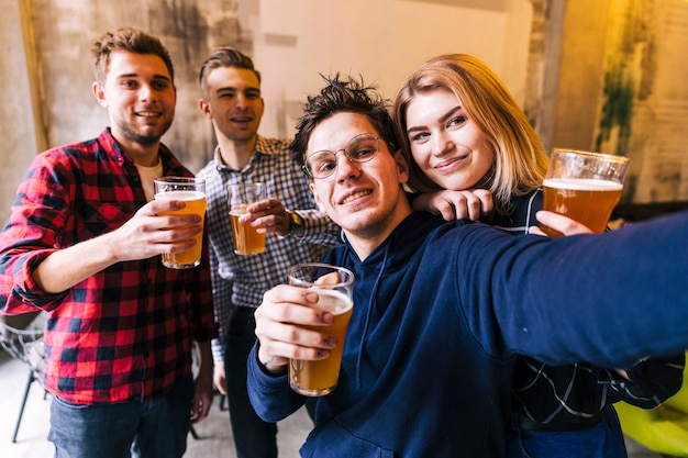 Pareja joven tomando selfie con sus amigos sosteniendo los vasos de cerveza