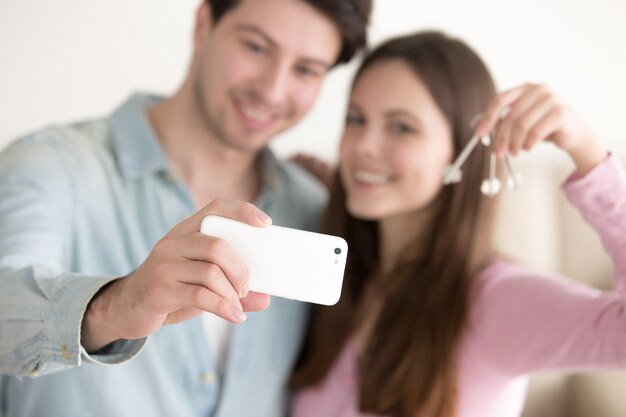 Pareja joven tomando selfie usando teléfono inteligente sosteniendo las llaves