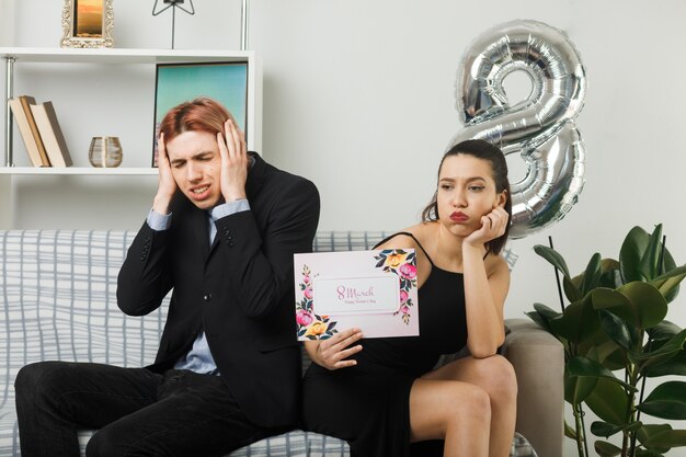 Pareja joven triste en el día de la mujer feliz con tarjeta de felicitación chico poniendo las manos en los oídos sentado en el sofá en la sala de estar