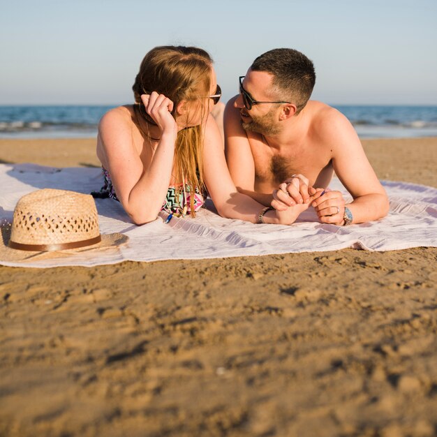 Pareja joven tumbado en una manta cerca de la orilla del mar mirando el uno al otro en la playa