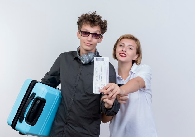 Pareja joven de turistas hombre con auriculares sosteniendo maleta mostrando billetes de avión de pie junto a su novia sonriendo confiado sobre la pared blanca