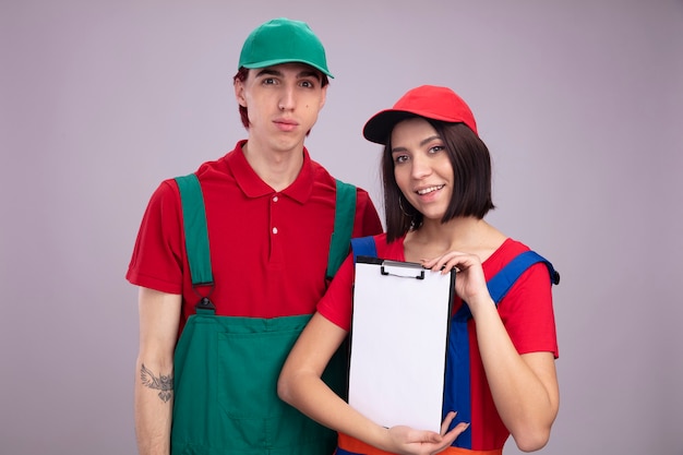 Pareja joven en uniforme de trabajador de la construcción y gorra chico serio chica sonriente tanto chica mostrando portapapeles