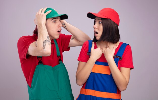 Pareja joven en uniforme de trabajador de la construcción y gorra mirando el uno al otro chico preocupado manteniendo las manos en la cabeza chica sorprendida apuntando a sí misma aislada en la pared blanca