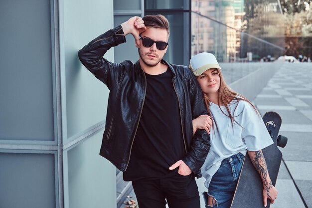 Pareja joven vestida con ropa de moda posando con patinetas cerca de rascacielos.