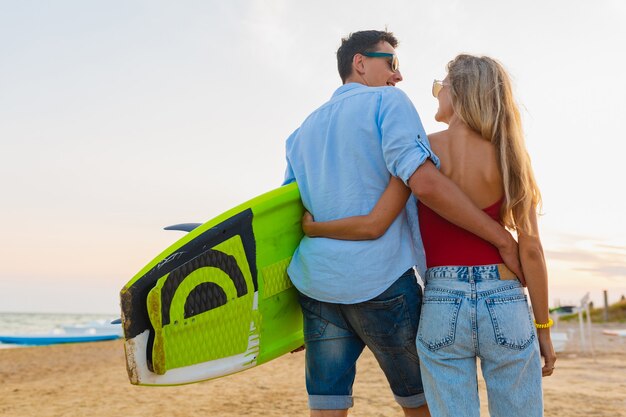 Pareja joven vista desde atrás divirtiéndose en la playa caminando con tabla de surf