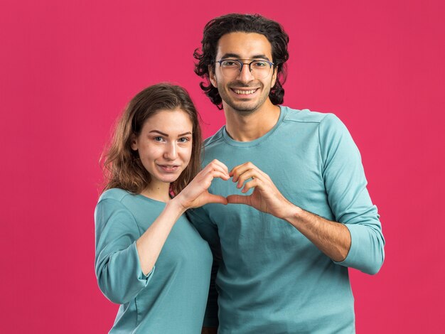 Pareja joven vistiendo pijamas hombre sonriente con gafas mujer complacida ambos mirando al frente haciendo signo de corazón juntos aislado en la pared rosa