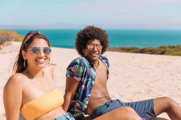 Pareja de jóvenes en gafas de sol sentado en la playa sonriendo y mirando a cámara