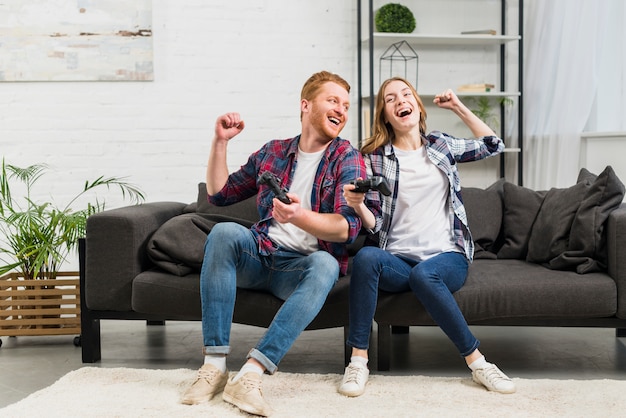 Pareja de jóvenes sentados en el sofá jugando al videojuego en la sala de estar