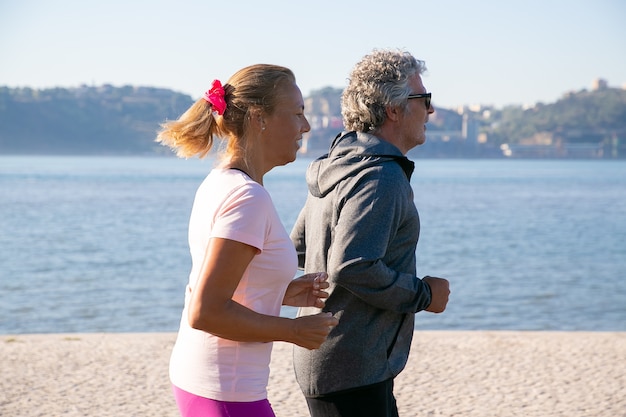 Foto gratuita pareja de jubilados vistiendo ropa deportiva, disfrutando de correr por la mañana, trotar a lo largo de la orilla del río por la mañana. vista lateral. concepto de estilo de vida y jubilación