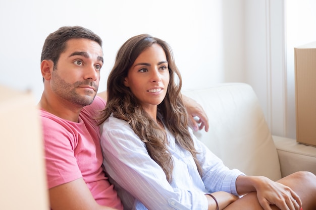 Pareja latina centrada en positivo sentado en el sofá entre cajas de cartón en el nuevo apartamento, mirando a otro lado