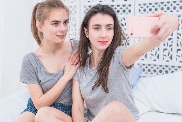 Pareja de lesbianas jóvenes sentados en la cama tomando selfie en el teléfono móvil