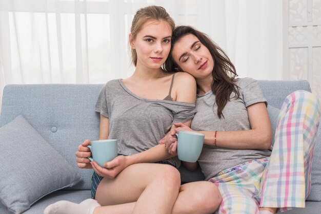 Pareja de lesbianas jóvenes sentados en un sofá sosteniendo una taza de café en la mano