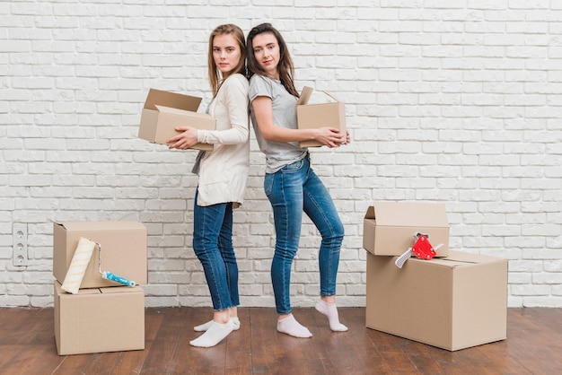 Foto gratuita pareja de lesbianas jóvenes sosteniendo cajas de cartón en movimiento en la mano de pie de espaldas contra la pared blanca