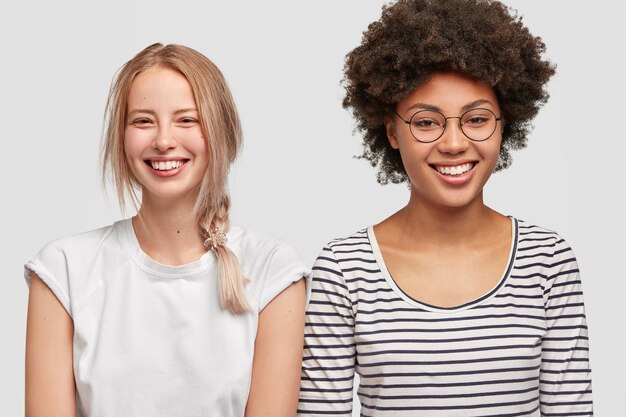 La pareja de lesbianas multirracial tiene expresiones positivas. Feliz joven rubia europea y su mejor amiga afroamericana, sonríen ampliamente, estando de buen humor. Concepto de amistad y emociones