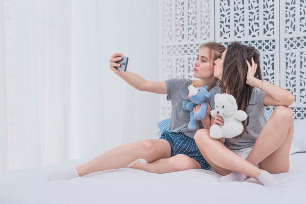 Pareja de lesbianas sentadas en la cama sosteniendo peluches tomando selfie en un teléfono móvil