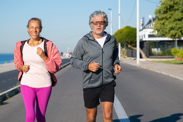 Pareja madura alegre a lo largo de la orilla del río. Hombre y mujer de pelo gris con ropa deportiva, trotar afuera. Concepto de edad y estilo de vida activo