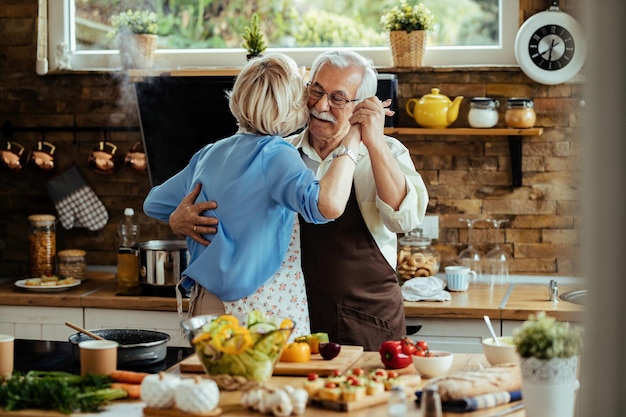 Pareja madura despreocupada bailando mientras prepara la comida en la cocina.