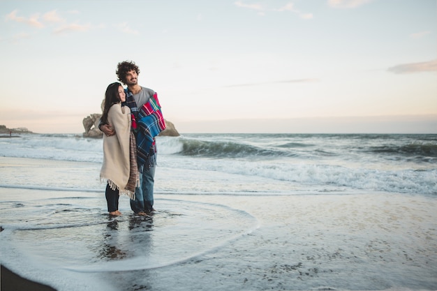 Foto gratuita pareja con mantas puestas encima y los pies en el agua de la playa