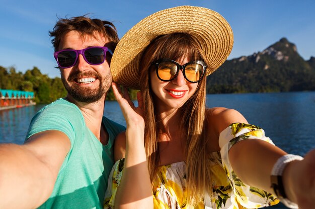 pareja marcando selfie cerca de la increíble vista del lago y las montañas, vistiendo ropa y accesorios elegantes. Ambiente alegre y juguetón.