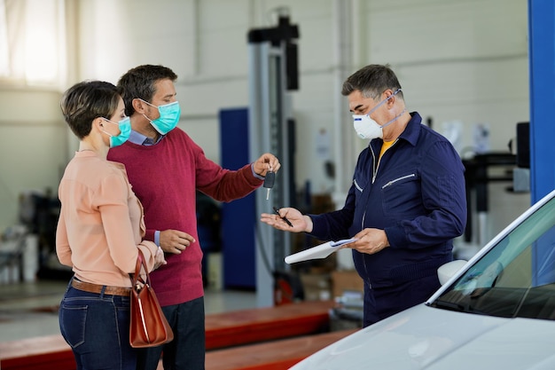 Pareja con mascarillas protectoras que dan la llave del auto a su reparador de automóviles en un taller