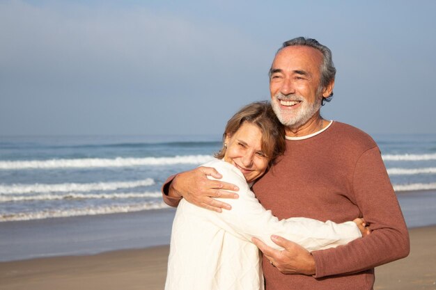 Pareja mayor caucásica pasando tiempo en la orilla del mar. Hombre canoso con barba sonriendo alegremente mientras abraza a su amada esposa. Fondo marino. Tiro medio. Relación, viaje, concepto de jubilación.