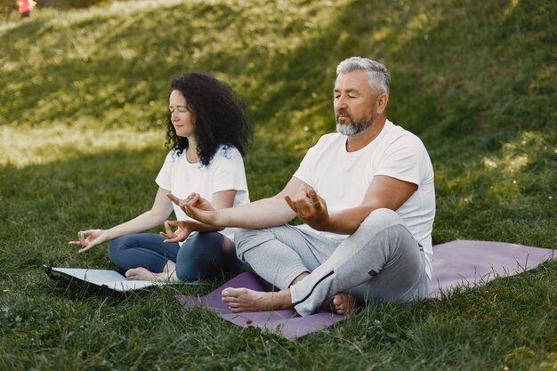 La pareja mayor está haciendo yoga al aire libre. Estirarse en el parque durante el amanecer. Morena con camiseta blanca.