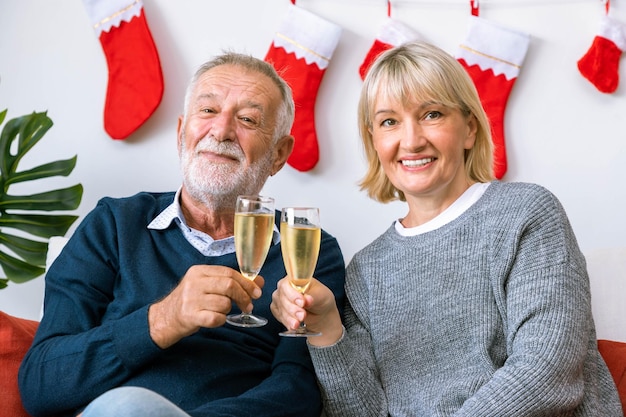 Pareja mayor hombre y mujer con champán y charlando sentados en un sofá con árbol de Navidad y decoración en segundo plano.