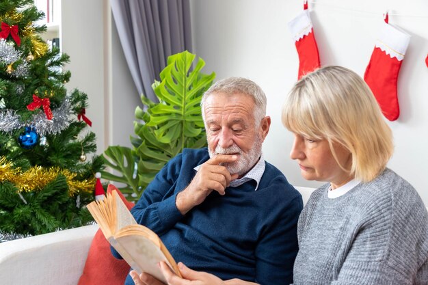 Pareja mayor hombre y mujer leyendo un libro sentado en un sofá con decoración de árbol de Navidad en segundo plano.