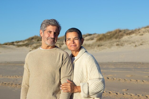 Una pareja mayor pasa tiempo afuera en la orilla del mar juntos, disfrutando del cálido sol de la tarde. Mujer de pelo corto sosteniendo la mano del hombre y sonriendo mientras un apuesto hombre barbudo mira la cámara. Concepto de relación