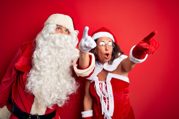 Una pareja de mediana edad con disfraz de Papá Noel y gafas sobre un fondo rojo aislado señalando con el dedo sorprendido hacia adelante boca abierta expresión asombrada algo en el frente