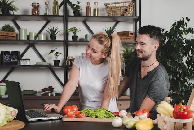 Pareja mirando la computadora portátil de pie detrás de la encimera con verduras