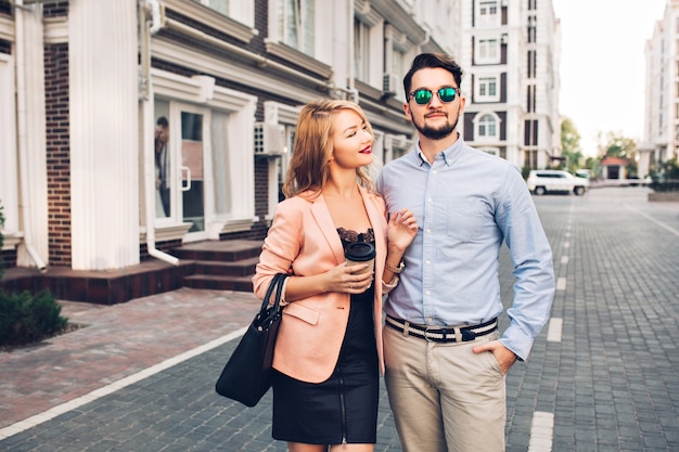 Pareja de moda está caminando por la calle en la ciudad. Chico guapo con barba en gafas de sol está abrazando a chica y mira lejos.