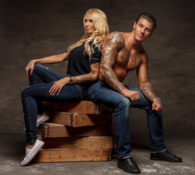 Pareja de moda moderna. Hombre tatuado sin camisa y sensual mujer rubia sentada en bloques de madera.