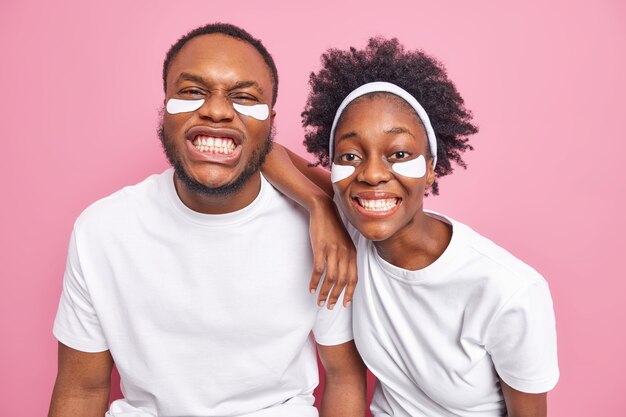 pareja muestra dientes blancos perfectos pasar tiempo juntos en tratamientos de belleza usar camisetas casuales aplicar almohadillas debajo de los ojos para hidratar aislado en la pared rosa