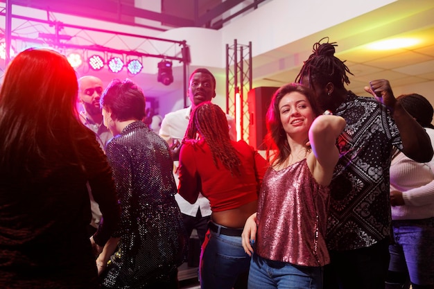 Foto gratuita pareja multirracial bailando juntos mientras festejaba en una discoteca llena de gente. hombre y mujer bailando y divirtiéndose en la pista de baile iluminada con focos en la discoteca del club