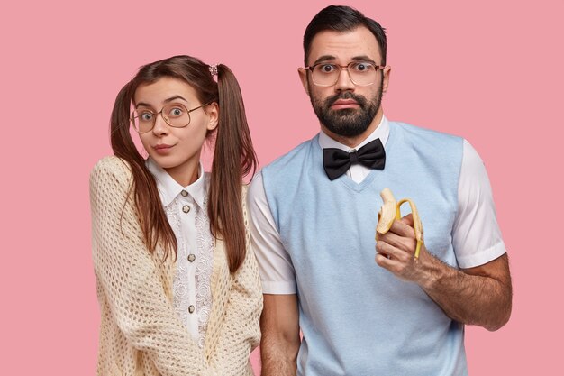 Pareja nerd vestida con traje de moda antiguo, grandes espectáculos, comer plátano, mirar confuso