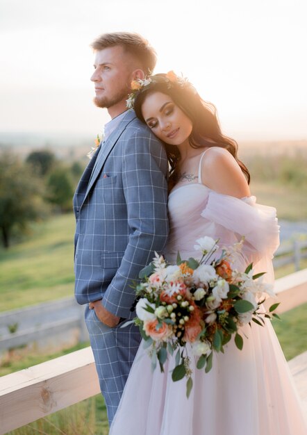 Pareja de novios en la cálida noche de verano cerca de la pradera vestida con un vestido de novia boho con hermoso ramo de novia